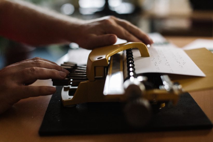 An einer Braille-Schreibmaschine wird gerade ein Dokument aufgesetzt. Man sieht lediglich das Gerät mit einem Stück Papier und die Hände des Nutzers.