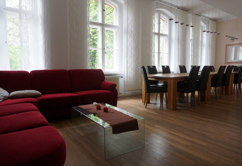 Ein Raum, in dem links eine rote Eckcouch steht und rechts eine Tischgruppe mit Stühlen.