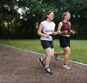 Zwei Frauen joggen nebeneinander auf einer Laufstrecke. Beide halten gemeinsam ein Band fest.
