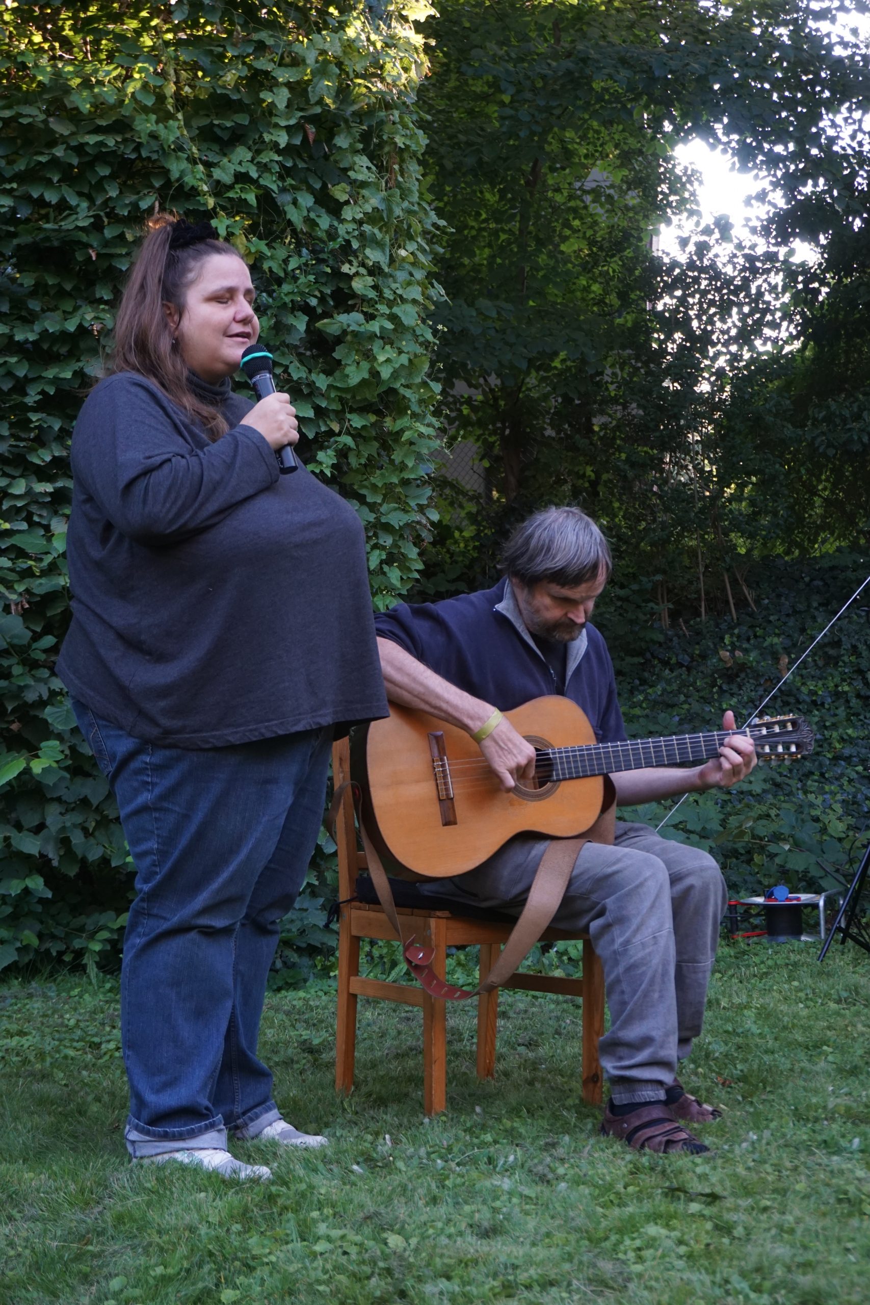 Eine Frau mittleren Alters steht neben einem sitzendem Gitarrenspieler. Beide sind draußen. Sie singt.
