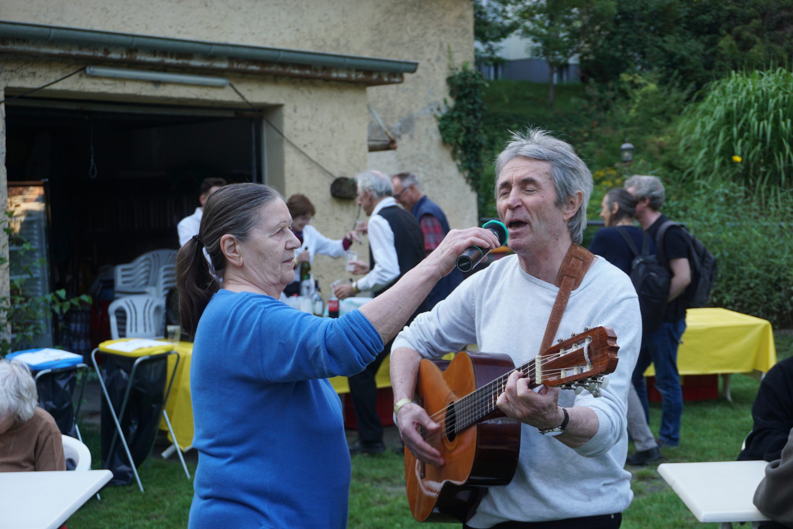 Ein Mann mit Gitarre singt. Eine Frau steht im Gegenüber und hält ein Mikrophon vor seinen Mund. Beide sind draußen. Im Hintergrund sind mehrere Menschen zu sehen.t