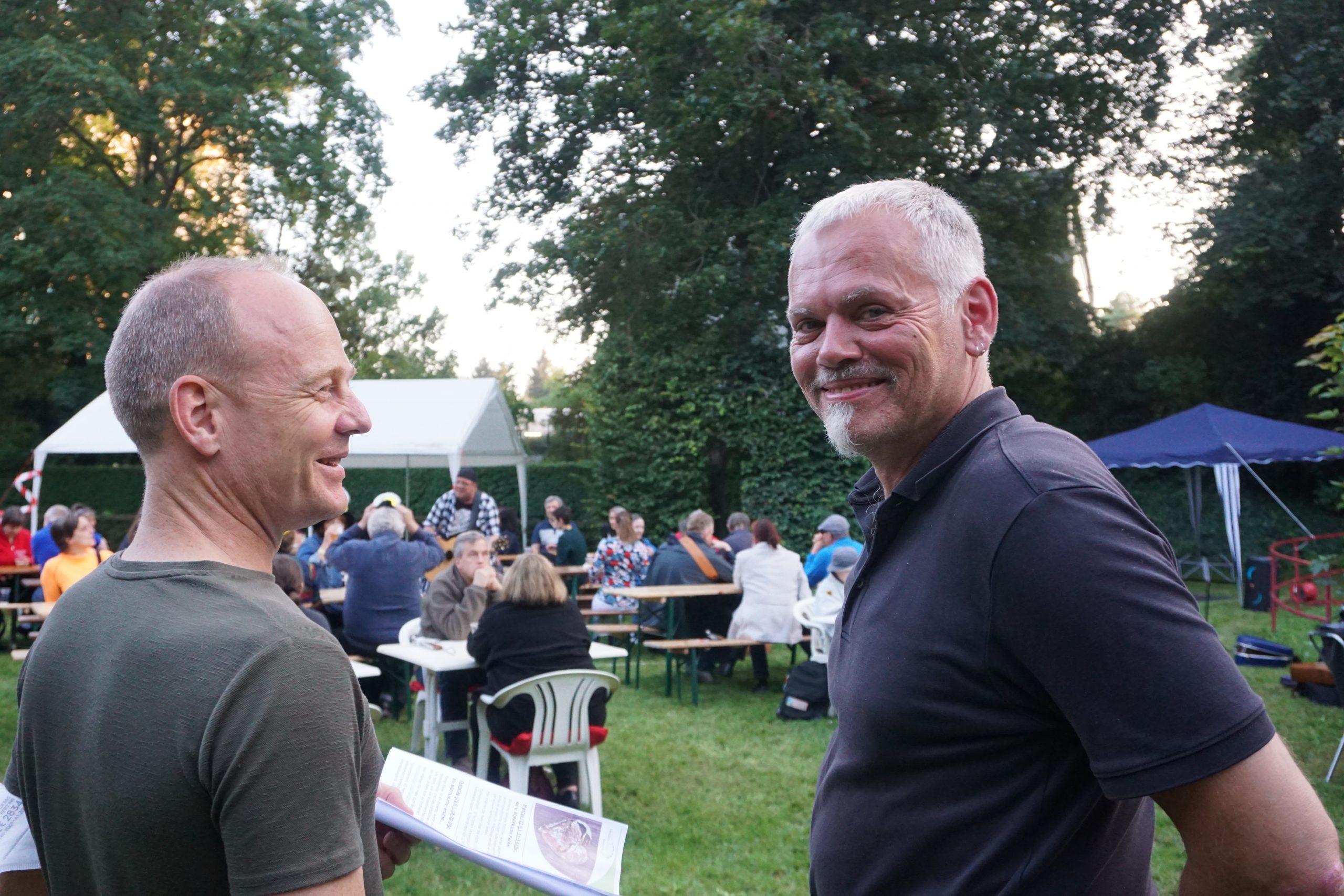 Herr Zehe (links) und Herr Ittner (rechts) stehen draußen und lächeln. im Hintergrund befinden sich Menschen, die an Bierbänken sitzen.