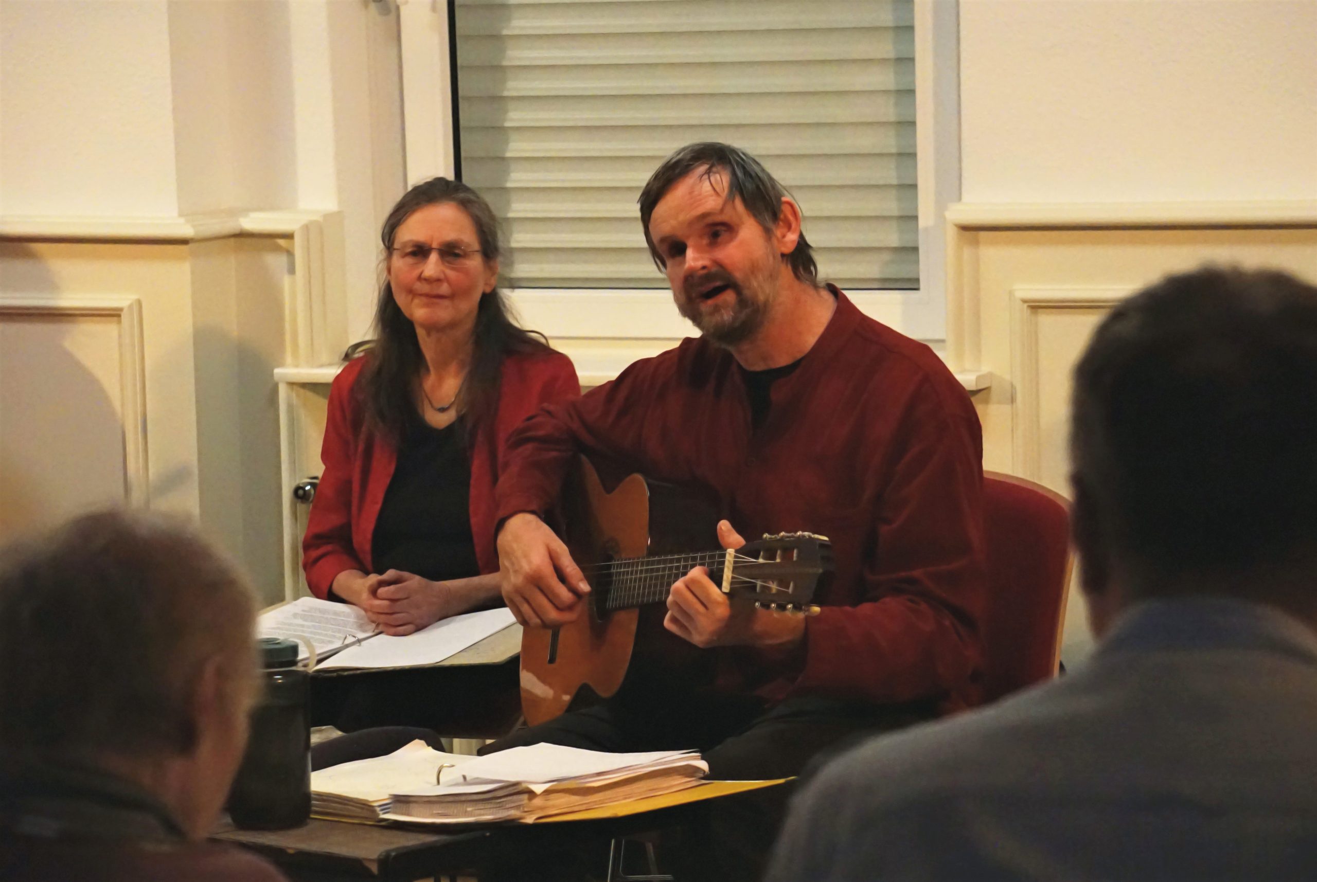 Ein Mann mit Gitarre sitzt spielend und singend neben einer Frau. Im Vordergrund erkennt man unscharfe Teile von Zuschauern.