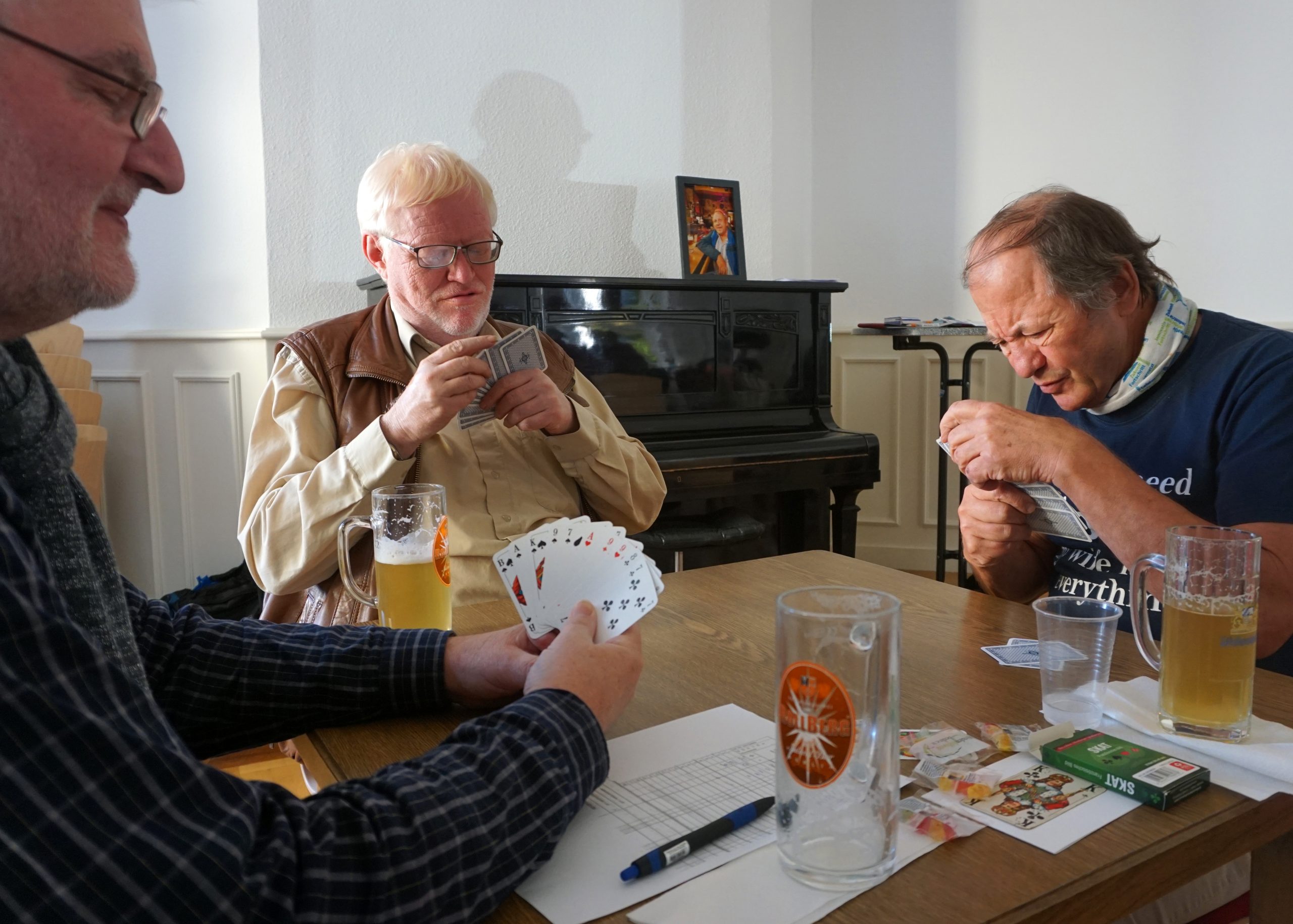 Drei Herren sitzen an einem Tisch und spielen Karten. Der Mann rechts auf dem Bild erkennt die Karten nur bei genauem Hinsehen.
