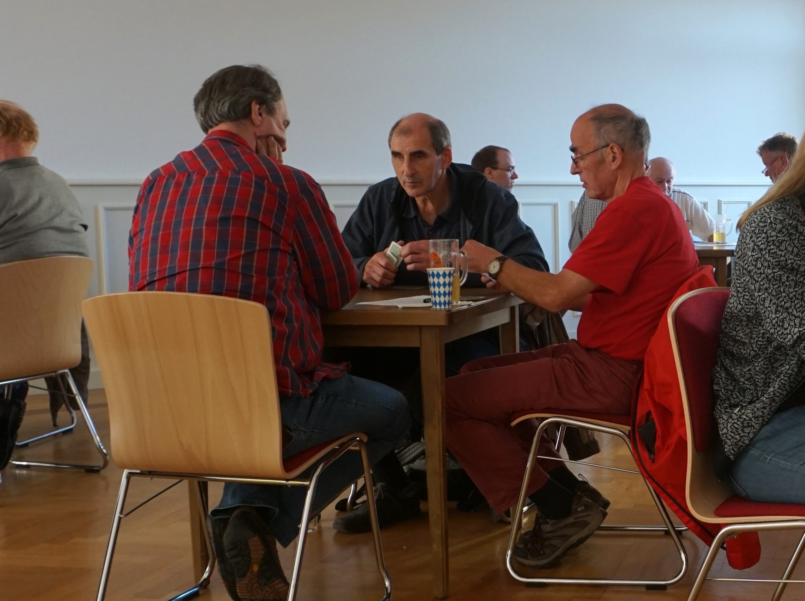Drei Männer sitzen an einem Tisch und spielen Karten. Ein Herr ist blind und erfühlt die Karten.