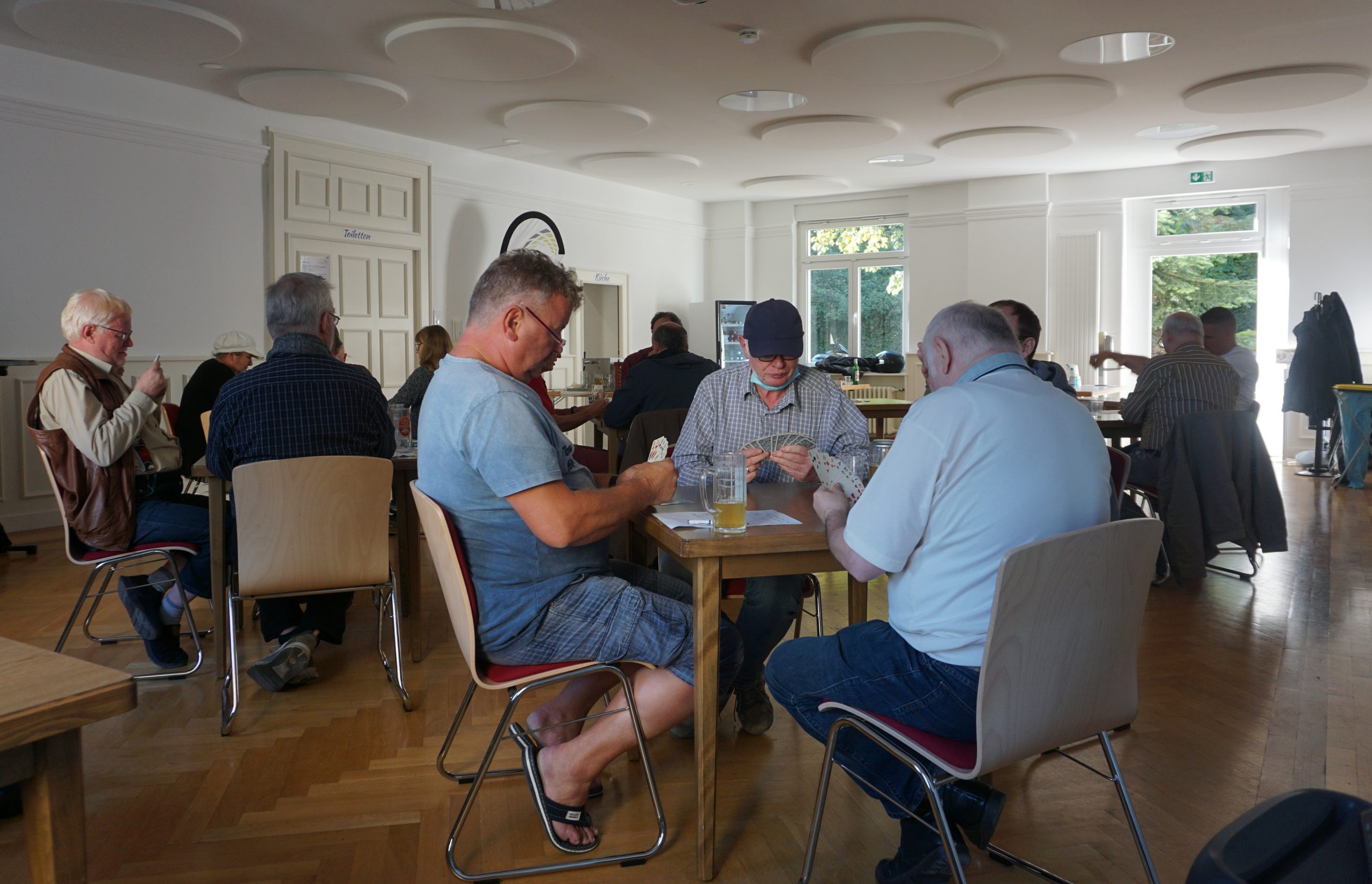 An mehreren Tischen spielen mehrere Personen in Dreiergruppen Karten.