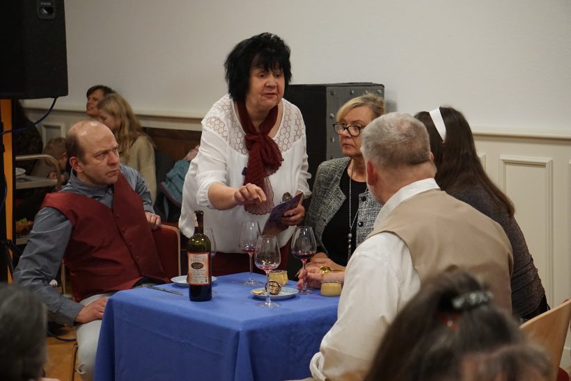 Ein Bild einer Theateraufführung. Zwei Damen und zwei Herren sitzen an einem Tisch. Vor jedem steht ein Glas Wein. Zwischen dem einen Herren und der einen Damen steht eine Dame mit weißer Bluse. Sie ist leicht über den Tisch gebeugt und sieht erklärend aus.