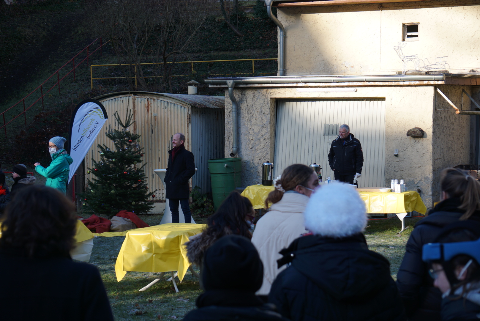 Ein Mann neben Weihnachtsbaum steht vor mehreren Menschen und hält eine Ansprache.