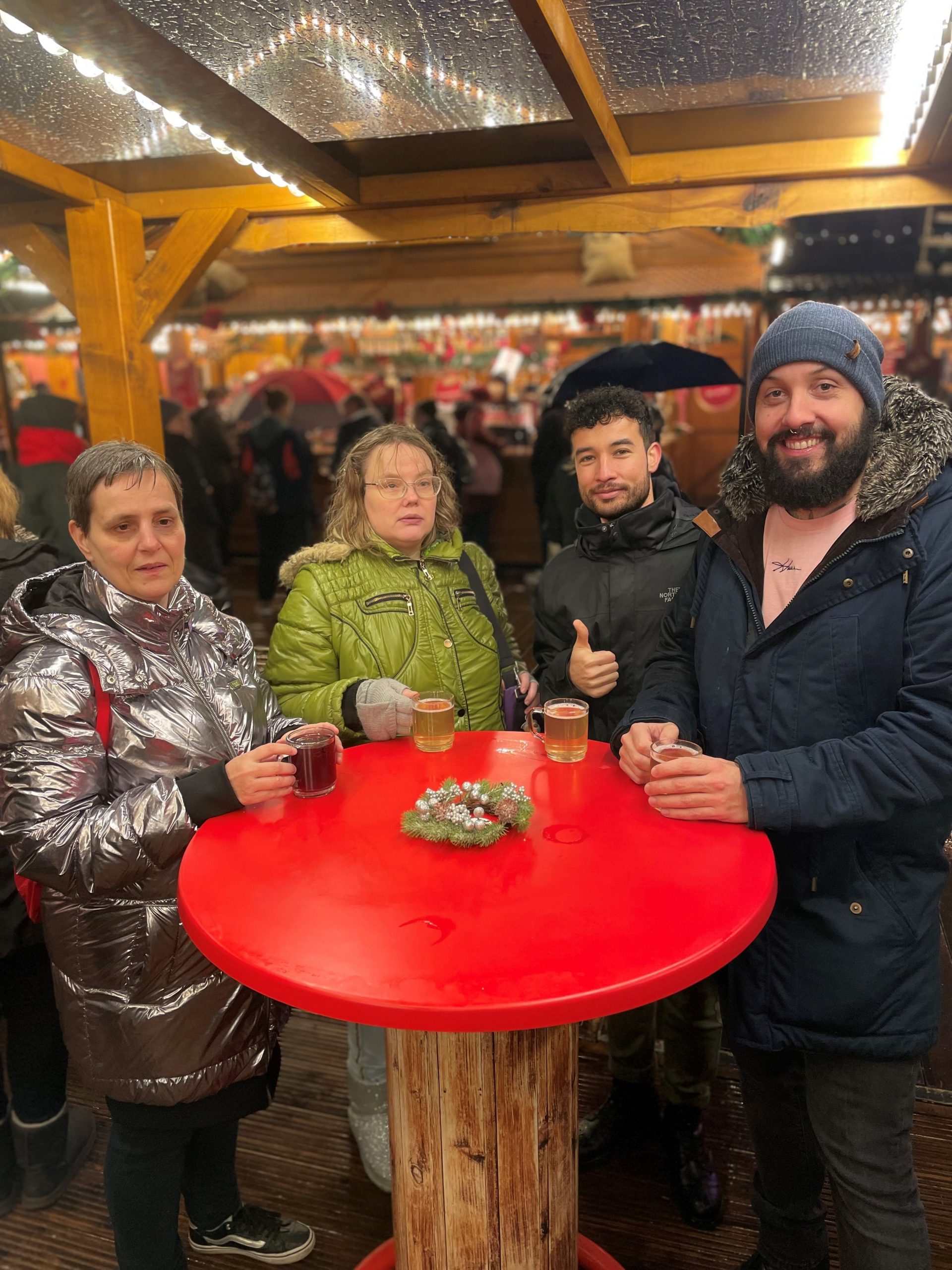 Zwei Frauen uns zwei Männer stehen in dicken Jacken an einem roten Stehtisch auf einem Weihnachtsmarkt. Alle haben ein Getränk vor sich zu stehen. 