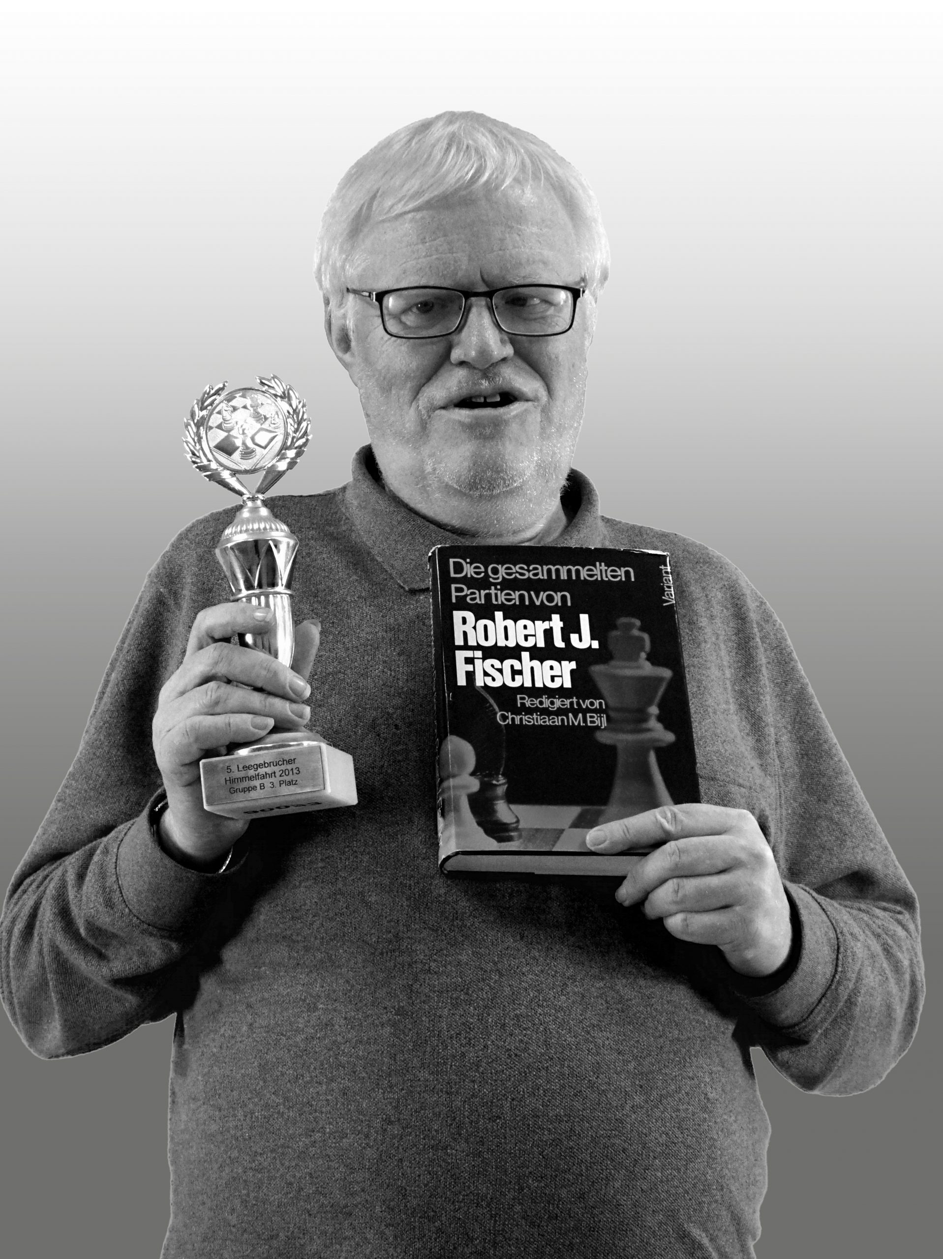 Hartmut Rakow blickt in die Kamera und lächelt. In der rechten Hand hält er einen Pokal, auf dessen Sockel „5. Leegebrucher Himmelfahrt 2013 Gruppe B 3. Platz steht“. In seiner linken Hand hält er ein Buch mit dem Titel „Die gesammelten Partien von Robert J. Fischer“.