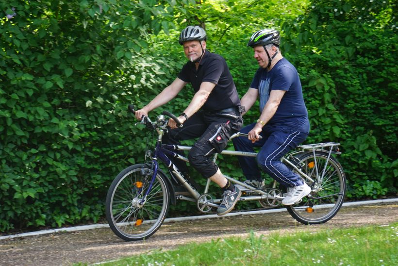 Zwei Männer mit Fahrradhelmen auf dem Kopf fahren gemeinsam Tandem.