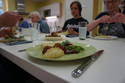 Ein belegter Teller steht auf einem Tisch, an dem mehrere Personen sitzen. Alle haben etwas zu essen vor sich zu stehen.