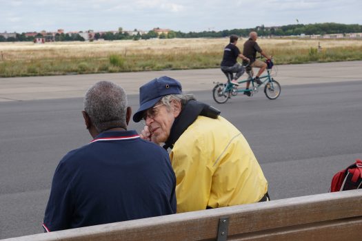 Zwei Herren sitzen auf einer Bank auf dem Tempelhofer Feld. Im Hintergrund fahren zwei Personen Tandem.