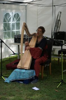 Eine Frau sitzt in einem Zelt. Sie spielt Harfe. Sie trägr ein keltisches Kleid und singt.