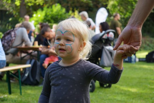 Ein Portraitbild eines zweijährigen Mädchens. Es ist im Gesicht mit einem Schmetterlingsmotiv geschmickt. Es hält die Hand einer größeren Person, die nicht zu sehen ist.