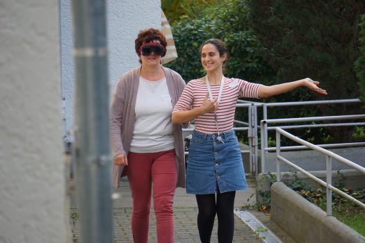 Zwei Damen laufen einen Weg entlang. Die Linke trägt eine Schweißerbrille und hält sich am Arm der anderen Frau fest. Diese streckt ihren linken Arm aus und lächelt.