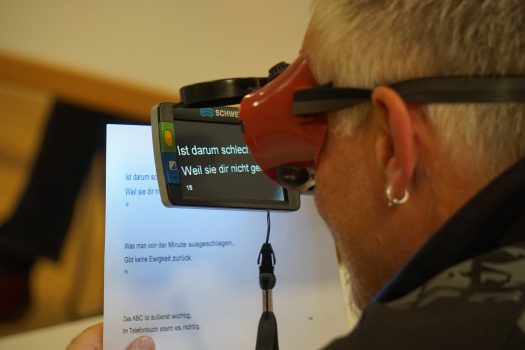 Ein Mann mit Schweißerbrille schaut durch eine elektronische Lupe, die die Farben invertiert, auf ein weißes Blatt Papier mit schwarzer Schrift.