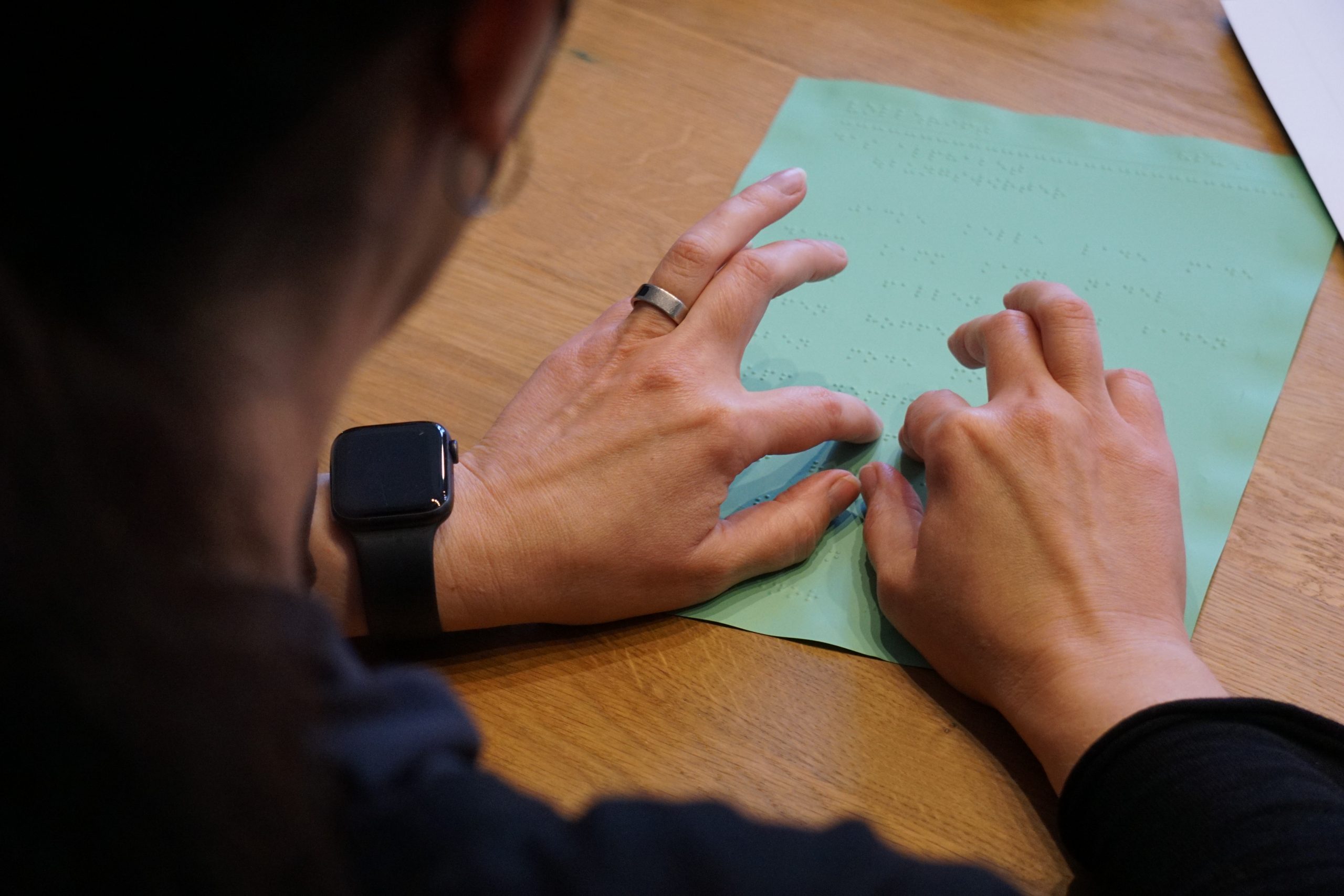 Das Bild blickt über die Schulter einer Frau. Sie liest einen grünen Zettel mit Punktschrift. Ihre Finger fahren gerade über das Blatt.