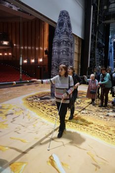 Eine Stange auf einer Bühne wird als Handlauf von blinden Menschen genutzt. Sie laufen über einen gelb gemusterten Teppich.