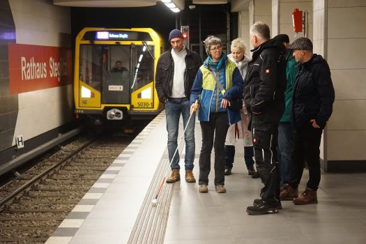 Eine Gruppe bestehend aus 6 Personen steht an einem U-Bahn-Gleis. Ein Zug fährt ein. Eine Frau hält einen Blindenstock.
