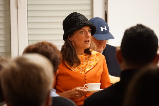 Eine Dame mittleren Alters mit Hut hält eine Kaffeetasse mit Untertasse und schaut entsetzt. Vor ihr sitzt Publikum.