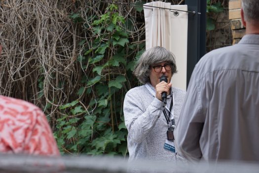 Ein Herr mittleren Alters mit wuscheligen grauen Haaren steht vor einigen Personen und spricht in ein Mikrophon.