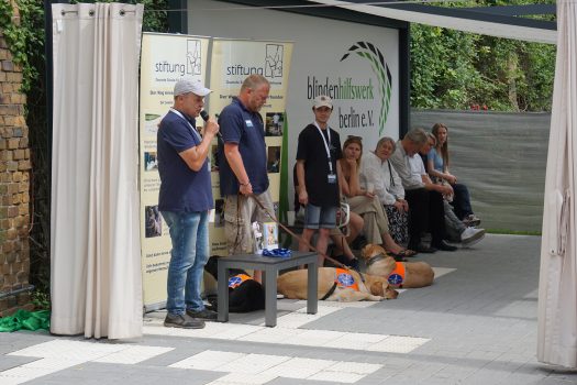 Drei Herren mit Blindenführhunde stehen vor zwei Roll-Ups und sprechen zu verschiedenen Menschen.
