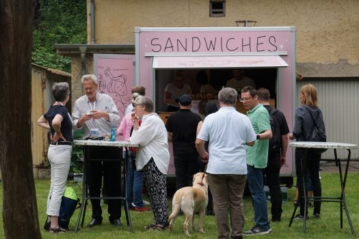 Mehrere Menschen stehen vor einem rosa Foodtruck, auf dem Sandwiches steht, an Stehtischen.